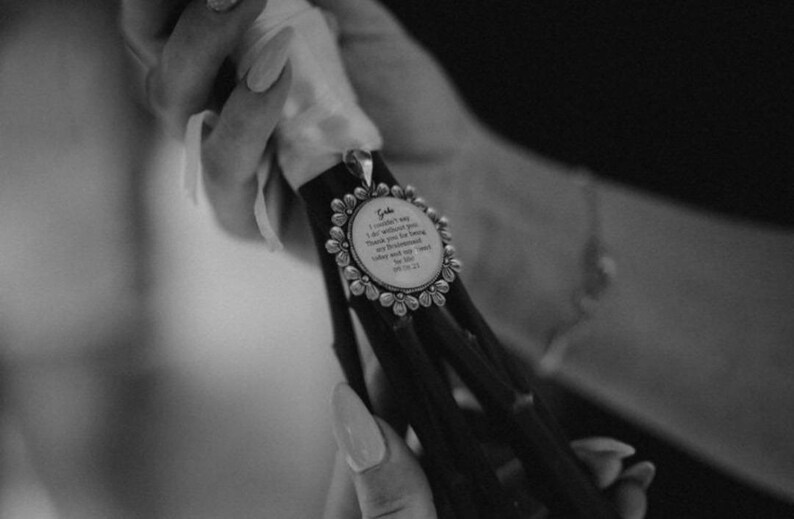 May include: A black and white image of a bouquet with a decorative pendant. The pendant is round with a floral border and contains text. The text reads: "Gabe I couldn't say I do without you. Thank you for being my bridesmaid..." and the date 06.26.21.