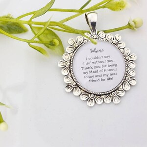 May include: A silver-toned pendant with a floral border. The pendant has a white center with the name "Sabine" and a message about being a Maid of Honor. The pendant is on a white surface with green foliage.