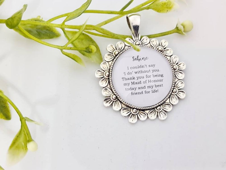 May include: A silver-toned pendant with a floral border. The pendant has a white center with the name "Sabine" and a message about being a Maid of Honor. The pendant is on a white surface with green foliage.