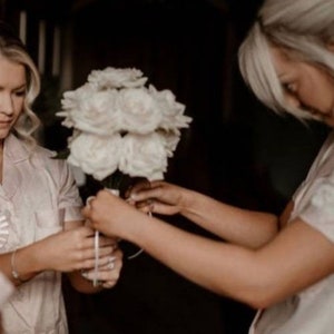 May include: A bouquet of white roses is being prepared. Two people are shown, one holding the flowers and the other tying a ribbon around the stems. The background is dark, with the focus on the floral arrangement.