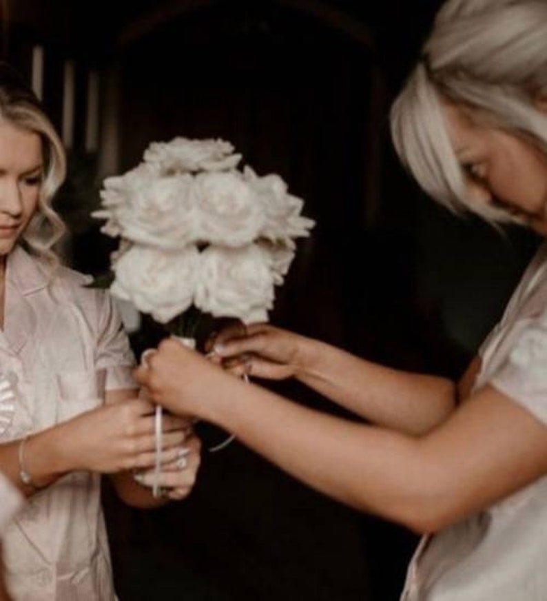 May include: A bouquet of white roses is being prepared. Two people are shown, one holding the flowers and the other tying a ribbon around the stems. The background is dark, with the focus on the floral arrangement.