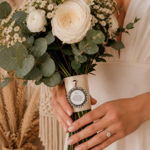 May include: A bridal bouquet featuring large, creamy white roses, small white flowers, and green foliage. The stems are wrapped with a beige ribbon, adorned with a silver pendant. The pendant has text. A diamond ring is visible.