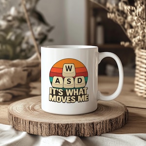 May include: White ceramic mug featuring a retro sunset design with the letters "W", "A", "S", and "D" in a keyboard key style. The text "IT'S WHAT MOVES ME" is below the keys. The mug is on a wooden surface.