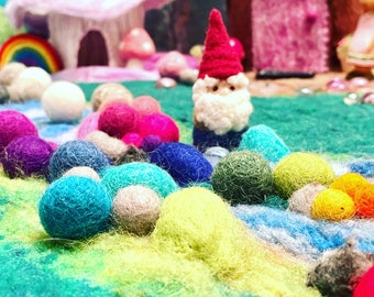 May include: A needle-felted gnome with a red pointed hat and white beard stands on a textured green landscape. The scene is decorated with numerous small, colorful felted wool balls in shades of blue, pink, yellow, and green, alongside miniature structures.