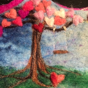 Puede incluir: Una obra de arte de fieltro que representa un árbol con un columpio colgando de sus ramas. El árbol está cubierto de corazones de fieltro rosa, rojo y blanco. El fondo es un cielo azul con nubes.
