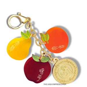 May include: A gold-colored keychain with a lobster clasp, featuring charms shaped like a yellow pear, a red apple, and an orange circle. Each charm has green leaves and gold accents. A gold coin with an embossed design is also attached.