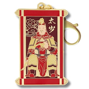 May include: A rectangular, red and gold pendant featuring a seated figure with a sword. The figure is dressed in ornate armor and holds a decorative object. The pendant has a gold clasp and a gold loop for attachment. The text "太岁" is visible.