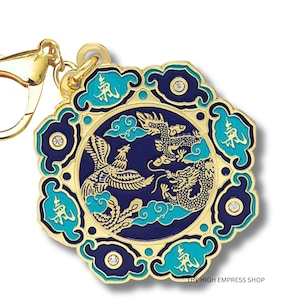 May include: A gold-colored keychain charm with a circular design featuring a phoenix and dragon in a blue and gold color scheme. The charm has a decorative border with turquoise accents and small crystals. The text "THE HIGH EMPRESS SHOP" is visible.