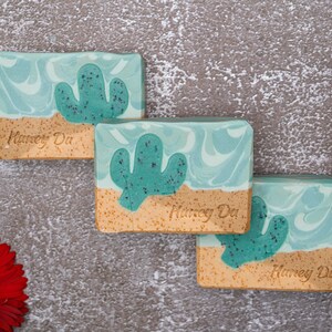 May include: Three bars of handmade soap with a cactus design. The soap is a light blue and green color with a brown sand-like base. The soap is labeled "Honey Du".