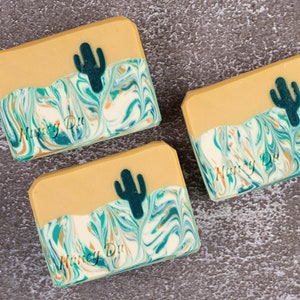 May include: Three bars of handmade soap with a desert theme. Each bar features a green and blue marbled design with a silhouette of a cactus on top. The soap is labeled "Nancy Du"