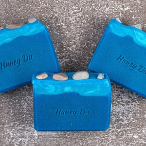 May include: Three handmade blue soaps with a wave design and small stones on top. The soaps have the text "Huney Du" embossed on them. The soaps are arranged on a textured surface with a yellow flower in the background.