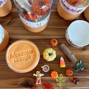 Peut inclure: Un pot rond de pâte orange avec le texte Montana dough en relief, entouré de breloques miniatures sur le thème de l'automne, dont des citrouilles, un épouvantail, une tasse de café, du maïs sucré, des feuilles, une brindille et une dinde.