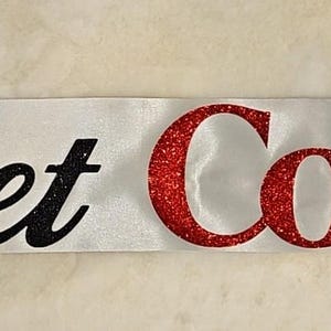 May include: A white satin sash with the words "Diet Coke" printed across it. The word "Diet" is in black glitter script, and "Coke" is in red glitter. The sash is rectangular with angled ends.