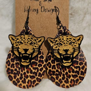May include: A pair of dangle earrings featuring a gold jaguar face on a brown and black leopard print background. The earrings are made of leather and have black metal hooks.