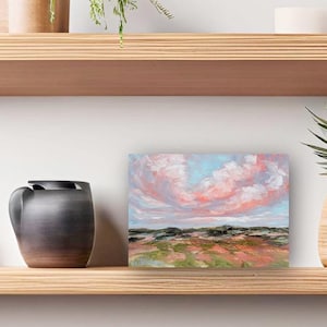 May include: A painting of a pink and blue sky with clouds over a green field. The painting is framed and hanging on a shelf.