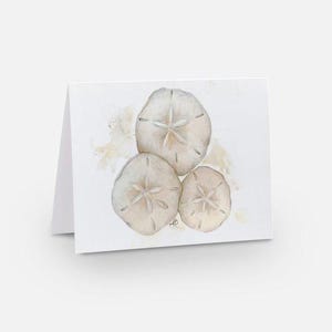 May include: A folded greeting card featuring a watercolor painting of three sand dollars in shades of beige and cream. The sand dollars are arranged in a cluster on a white background, with subtle brushstrokes of color.
