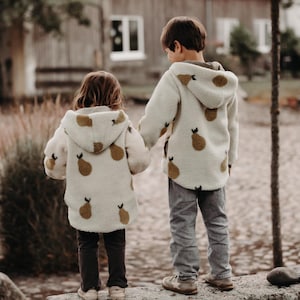 Könnte beinhalten: Zwei Kinder tragen cremefarbene Kapuzenjacken mit einem sich wiederholenden braunen Birnenmuster. Die Jacken sind mit grauen und braunen Hosen kombiniert. Sie stehen auf einer Steinoberfläche und halten Händchen, mit einem verschwommenen Hintergrund aus einem Gebäude und Bäumen.