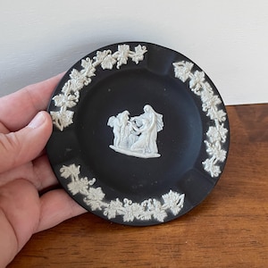 May include: Black ashtray with a white relief design. The ashtray features a white relief of a classical scene in the center and a white vine and leaf border. The ashtray is round and appears to be made of ceramic.