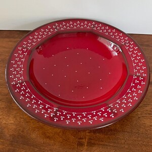 May include: A round, red ceramic plate with a glossy finish. The plate features a white dotted pattern in the center and a decorative border of small white floral designs. The plate is set on a wooden surface.