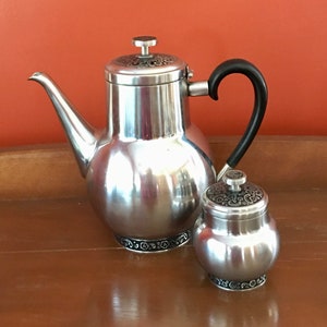 May include: A silver teapot and creamer set with black handles and ornate designs. The teapot has a round body and a spout. The creamer is a smaller, round container with a lid.