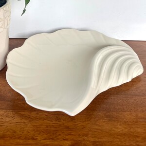 May include: White ceramic seashell dish with a wavy, textured design. The dish is shaped like a large seashell with a curved opening.