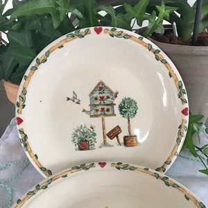 May include: Two white ceramic plates with a green leafy border and red hearts. The center of the plates features a birdhouse with a blue roof and green trim. There are two small birds flying around the birdhouse.