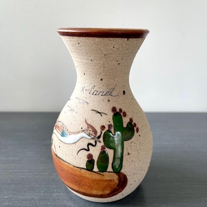 May include: A speckled ceramic vase with a brown rim and base. The vase features a hand-painted desert scene with a roadrunner, cactus, and the text "X-caret". The vase has a narrow neck and a rounded body.