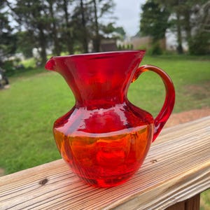 May include: A vibrant red glass pitcher with a handle and spout. The pitcher has a rounded body and a flared top. The glass appears to be transparent, with a slight orange hue at the base. The pitcher is sitting on a wooden surface.