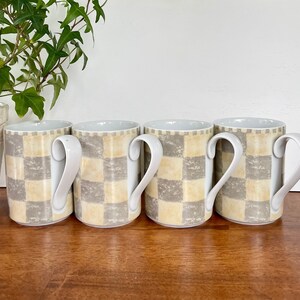 May include: Four white ceramic mugs with a checkered pattern in shades of beige and gray. The mugs have a rounded handle and are arranged in a row.