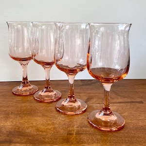 May include: Four pink-tinted glass goblets with a tulip-shaped bowl and a slender stem. The goblets are arranged on a wooden surface, showcasing their delicate design and color. The base of each goblet is round.