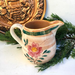 May include: A white ceramic pitcher with a green and brown striped band around the top. The pitcher has a floral design with a pink flower and green leaves.