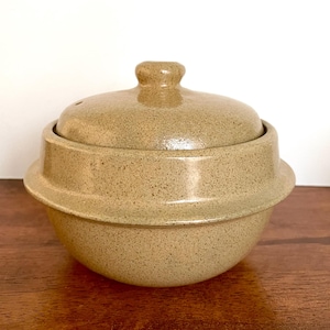 May include: A speckled tan ceramic cooking pot with a matching lid. The pot has a rounded base and a flared rim. The lid has a small knob on top for easy handling. The pot is designed for cooking and serving.