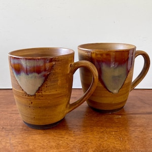 May include: Two handmade ceramic mugs with a rustic, earthy aesthetic. The mugs feature a warm brown base with a gradient of reddish-brown and cream-colored accents. Each mug has a curved handle and a dark base.