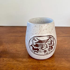May include: White ceramic mug with a brown speckled glaze. The mug features a brown silhouette of a horse-drawn wagon in front of a barn.