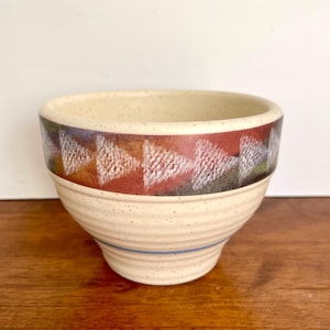 May include: A beige ceramic bowl with a geometric design. The rim features a band of red, orange, and purple triangles with white accents. A blue stripe encircles the base. The bowl has a speckled texture.
