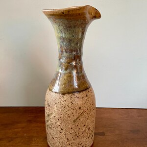May include: A brown and tan ceramic pitcher with a speckled glaze. The pitcher has a wide mouth and a narrow neck.