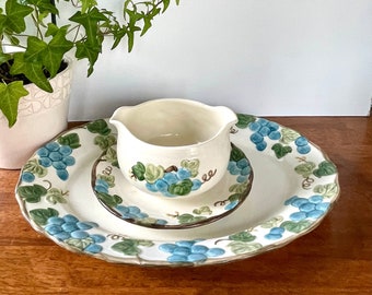 May include: A white ceramic gravy boat with a green and blue grape design sits on a matching white ceramic plate with a scalloped edge. The plate has a smaller plate inside it, also with a grape design.