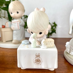 Puede incluir: Un conjunto de figuritas de porcelana de dos ni&ntilde;os, un ni&ntilde;o y una ni&ntilde;a, con cabello rubio. La ni&ntilde;a sostiene una mu&ntilde;eca peque&ntilde;a y una caja de regalo. El ni&ntilde;o sostiene un libro y una manzana roja. Las figuritas est&aacute;n sentadas en una mesa blanca con el logotipo de Precious Moments.