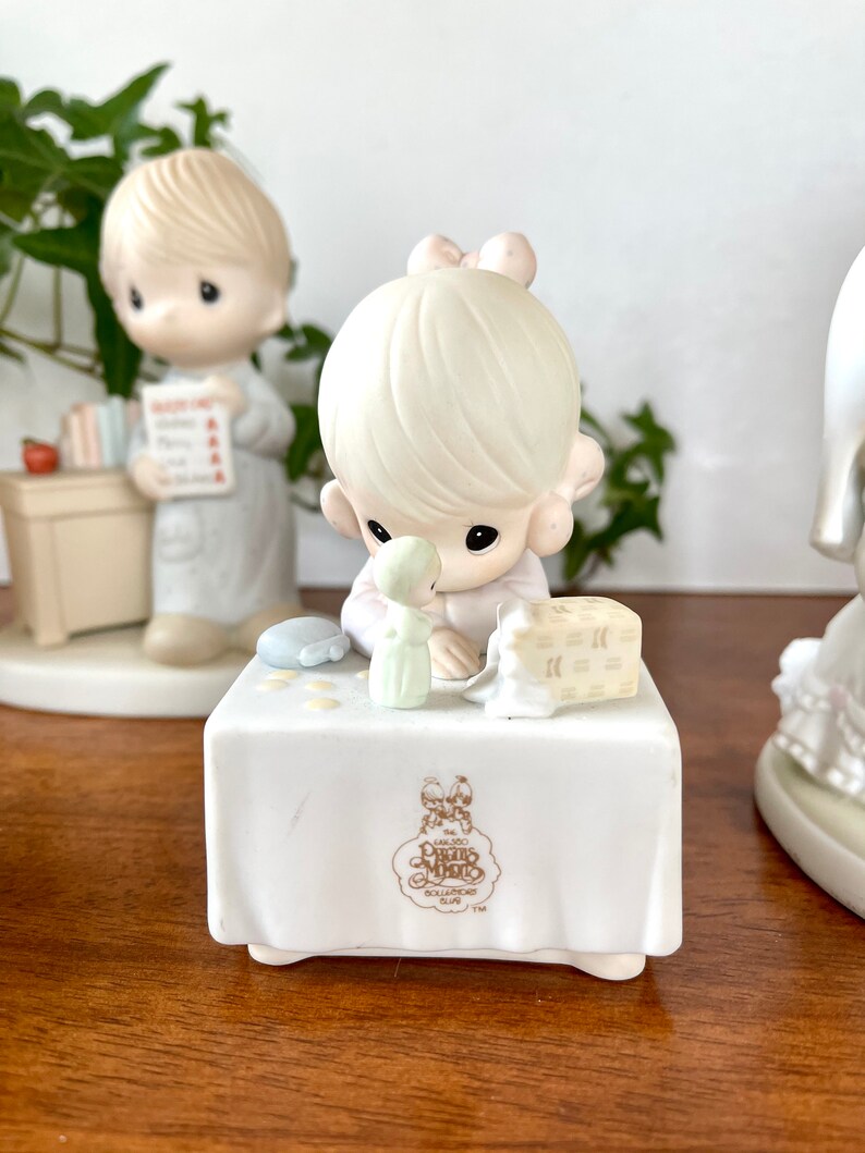 Puede incluir: Un conjunto de figuritas de porcelana de dos ni&ntilde;os, un ni&ntilde;o y una ni&ntilde;a, con cabello rubio. La ni&ntilde;a sostiene una mu&ntilde;eca peque&ntilde;a y una caja de regalo. El ni&ntilde;o sostiene un libro y una manzana roja. Las figuritas est&aacute;n sentadas en una mesa blanca con el logotipo de Precious Moments.