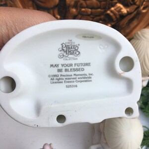 Puede incluir: Una figurita de porcelana blanca de una ni&ntilde;a que sostiene un ramo de flores. La figurita es parte de la colecci&oacute;n Precious Moments y tiene la inscripci&oacute;n "Que tu futuro sea bendecido".