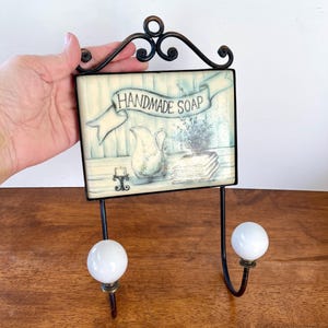 May include: A decorative wall hook with a rectangular sign that reads "HANDMADE SOAP." The sign features a watercolor-style illustration of a pitcher, flowers, and books. The hook has two white ball-shaped knobs and a black metal frame.