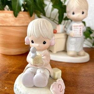Puede incluir: Una figurita de porcelana de una ni&ntilde;a peque&ntilde;a que sostiene un bloque con el n&uacute;mero "2" en &eacute;l. Est&aacute; sentada sobre una base con una decoraci&oacute;n en forma de coraz&oacute;n que tambi&eacute;n tiene el n&uacute;mero "2". La figurita es parte de una colecci&oacute;n Precious Moments.