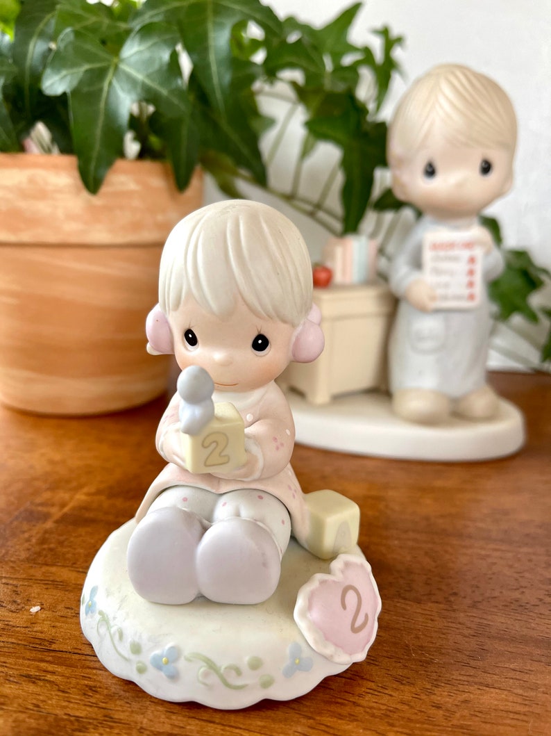 Puede incluir: Una figurita de porcelana de una ni&ntilde;a peque&ntilde;a que sostiene un bloque con el n&uacute;mero "2" en &eacute;l. Est&aacute; sentada sobre una base con una decoraci&oacute;n en forma de coraz&oacute;n que tambi&eacute;n tiene el n&uacute;mero "2". La figurita es parte de una colecci&oacute;n Precious Moments.