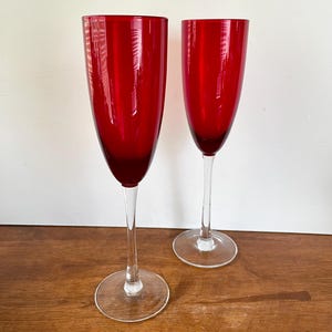 May include: Two tall champagne flutes featuring deep red glass bowls and clear glass stems and circular bases. The glasses have a slender, elongated shape designed for sparkling beverages, with a smooth, polished finish on the red glass surface.