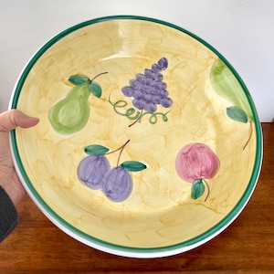 May include: Large, round decorative serving bowl with a yellow interior and a green rim. The bowl features hand-painted illustrations of fruit, including pears, grapes, plums, and a pink fruit. The bowl is likely made of ceramic or porcelain.