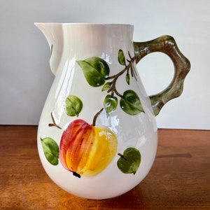 May include: White ceramic pitcher with a brown handle and a painted design of a red and yellow apple with green leaves.