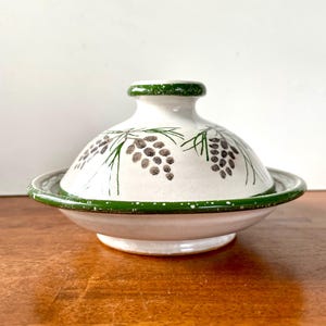 May include: A white ceramic serving dish with a lid. The lid and rim are decorated with green accents and a pine cone design. The dish is on a wooden surface.