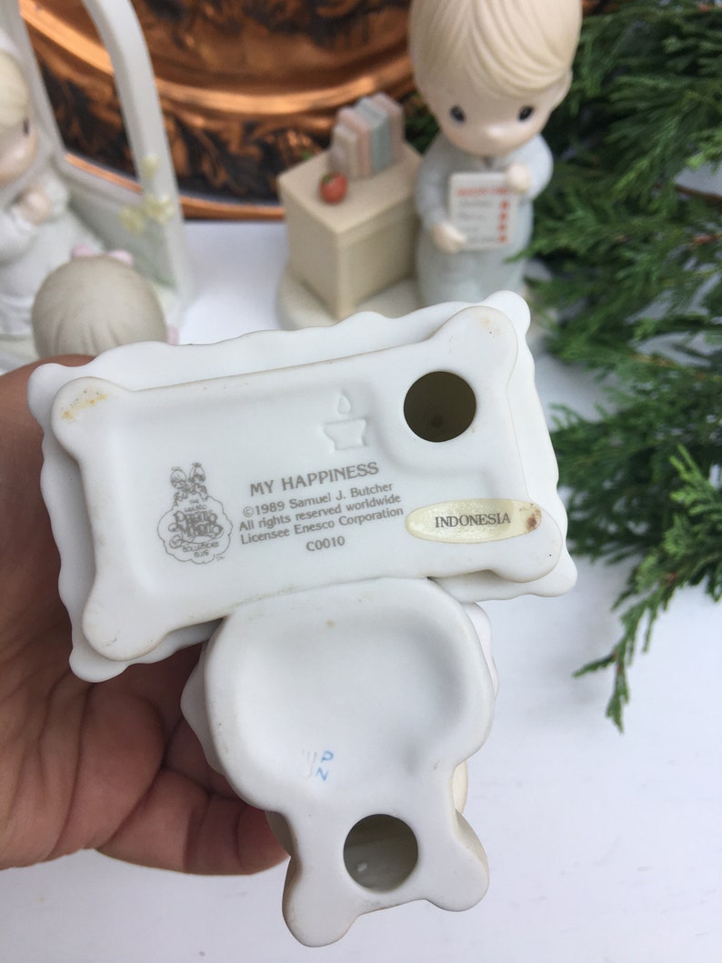 Puede incluir: Figurilla de porcelana blanca de una ni&ntilde;a sosteniendo una almohada con las palabras "My Happiness" en ella. La figurilla est&aacute; hecha por Precious Moments y est&aacute; marcada con "C0010" e "Indonesia".