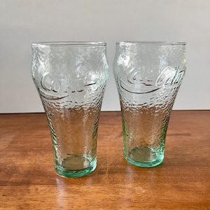 May include: Two clear Coca-Cola glasses with a textured surface. The glasses have a green tint at the base and feature the Coca-Cola logo. The glasses are displayed on a wooden surface.
