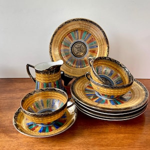 May include: A vintage tea set with gold rims and colorful geometric patterns. The set includes teacups, saucers, plates, and a creamer. The design features a central medallion with radiating stripes in shades of blue, red, green, and yellow.